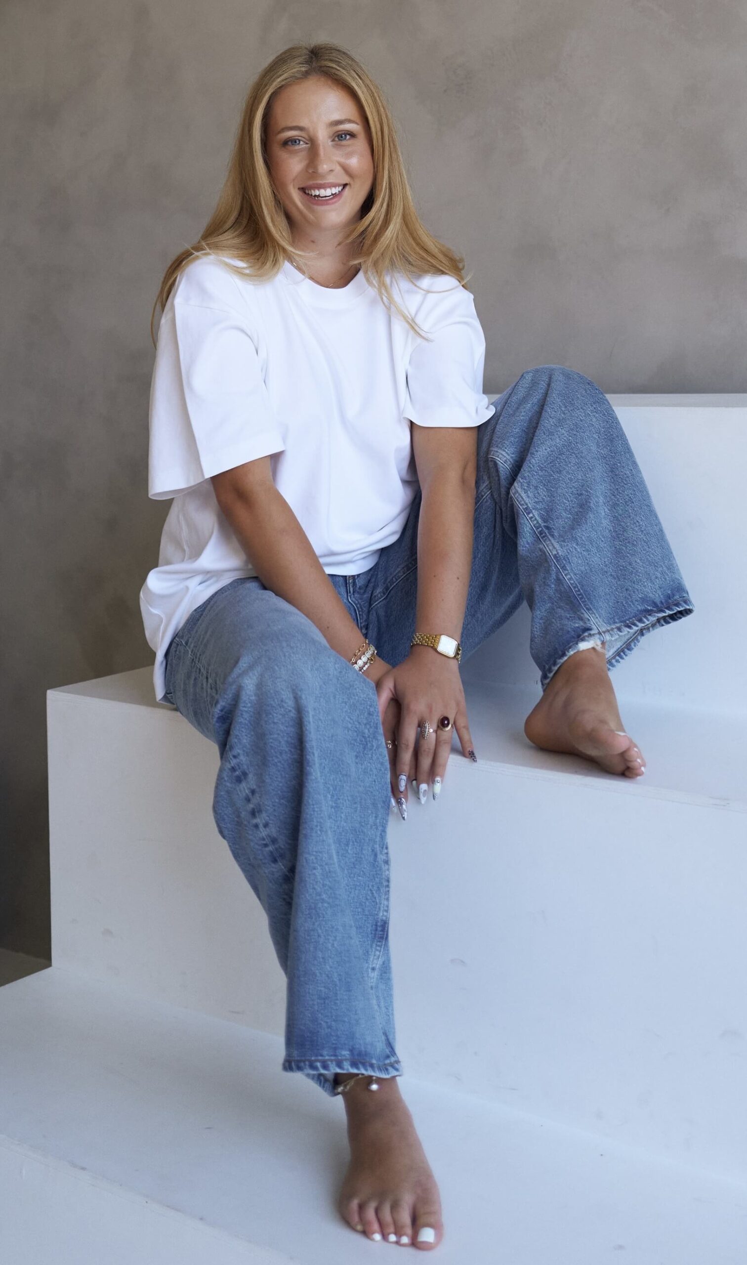 The Kind Kult A woman with long blonde hair sits barefoot on white steps, wearing a white t-shirt and blue jeans, smiling at the camera, radiating the peaceful vibe of mindfulness. Kind To Yourself. Kind To Others.