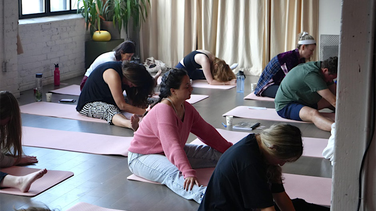 The Kind Kult People are seated on yoga mats in a studio, stretching forward with heads down. The room is softly lit with natural light, and there are plants and water bottles visible. Kind To Yourself. Kind To Others.
