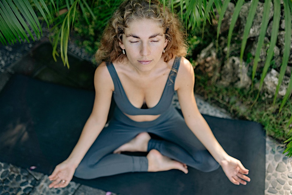 The Kind Kult Woman with curly hair sitting cross-legged on a yoga mat outdoors, eyes closed, practicing mindfulness with hands resting on knees—embracing peace and stillness as part of a supportive wellness community. Kind To Yourself. Kind To Others.