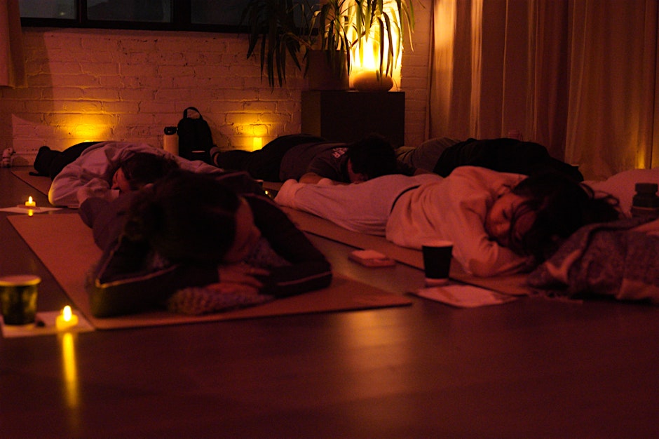 The Kind Kult Several people lie on yoga mats in a dimly lit room with warm lighting and candles, appearing to be resting or meditating. Kind To Yourself. Kind To Others.