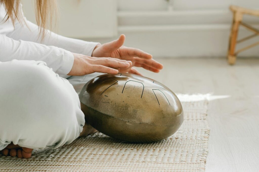 The Kind Kult A person in white sits cross-legged, practicing mindfulness as they play a metallic tongue drum with their hands on a woven mat in a bright room—a peaceful scene embraced by the wellness community. Kind To Yourself. Kind To Others.