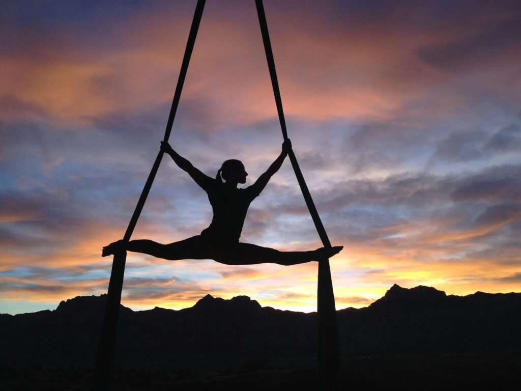 The Kind Kult A person practices mindfulness while performing a split on aerial silks, silhouetted against a colorful sunset sky and mountain landscape. Kind To Yourself. Kind To Others.