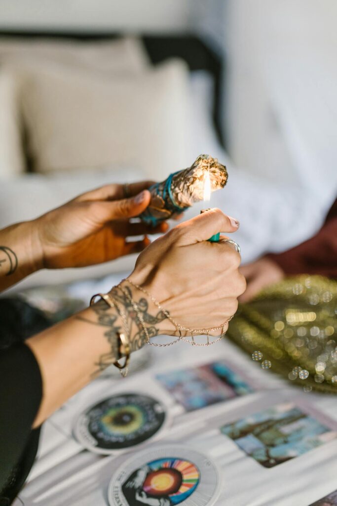 The Kind Kult A person lights a bundle of sage over a table with tarot cards and a golden mesh bag, wearing bracelets and rings—embracing mindfulness rituals often celebrated within the wellness community. Kind To Yourself. Kind To Others.