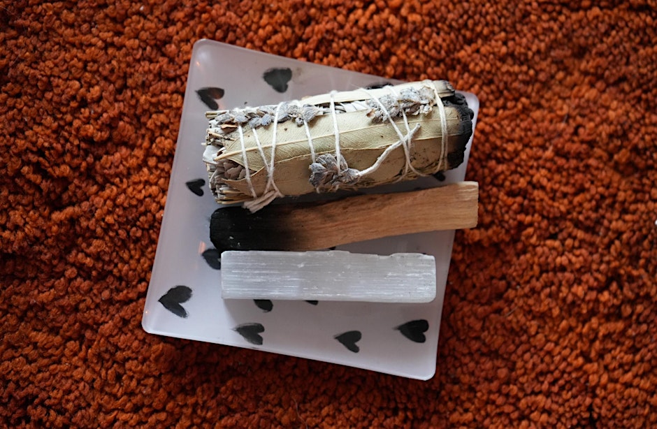 The Kind Kult A bundle of sage, a palo santo stick, and a selenite crystal rest on a small square dish with heart patterns, placed on an orange textured surface. Kind To Yourself. Kind To Others.