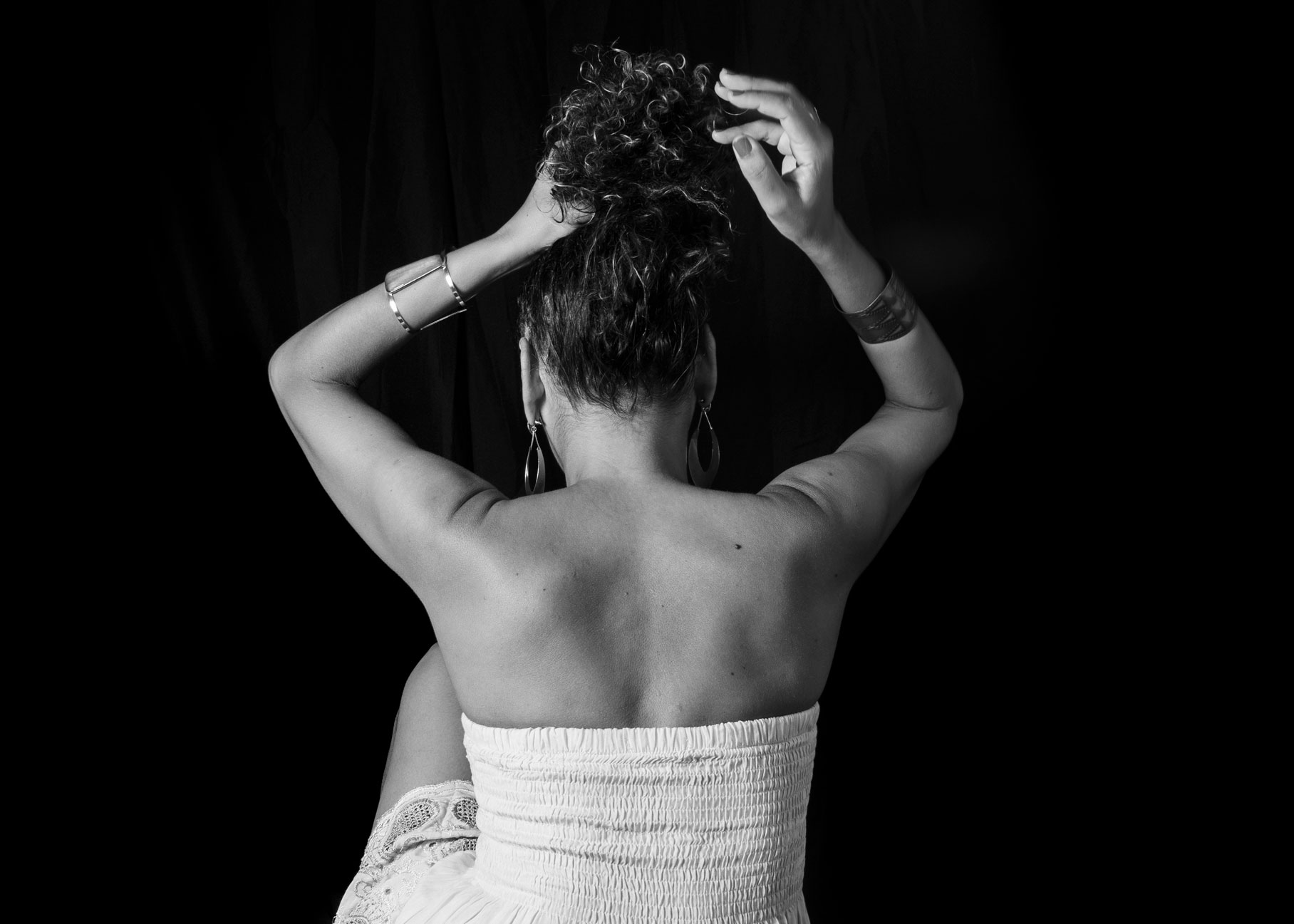 The Kind Kult A woman in a strapless dress sits with her back to the camera, raising her arms to adjust her curly hair against a black background. Kind To Yourself. Kind To Others.