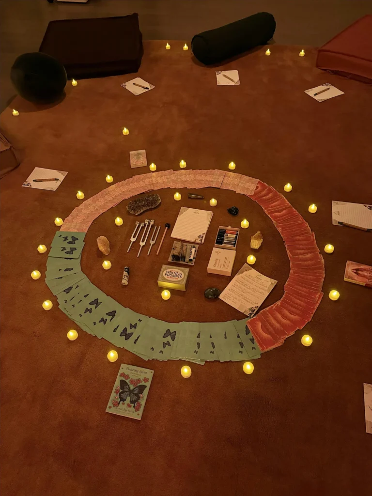 The Kind Kult A circular arrangement of cards, stones, and art supplies is surrounded by small LED candles on a brown carpet, with notebooks and cushions nearby. Kind To Yourself. Kind To Others.