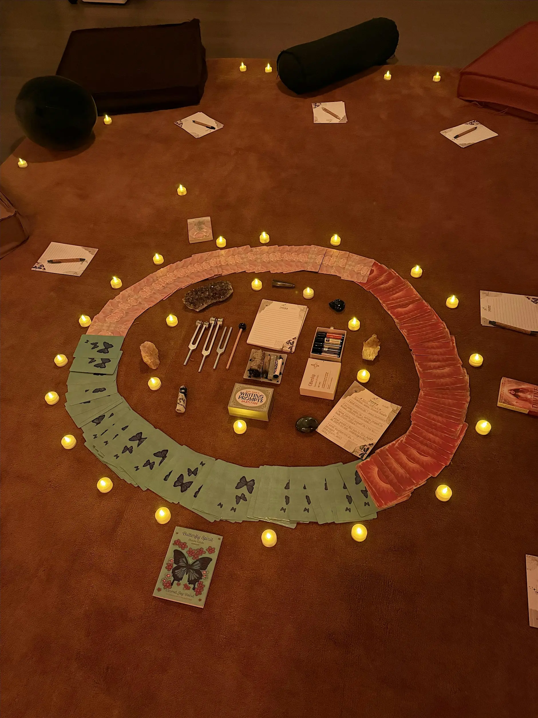 The Kind Kult A circular arrangement of cards, stones, and art supplies is surrounded by small LED candles on a brown carpet, with notebooks and cushions nearby. Kind To Yourself. Kind To Others.